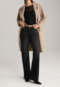 Béžový vlněný kabát přes černý vršek s vzorovanou průsvitnou látkou, černé džíny s vysokým pasem, béžový pásek s kulatou sponou a černé boty na podpatku se špičatou špičkou.