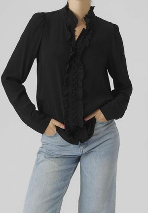 Žena v černé blůze s dlouhými rukávy, ozdobenou našitými volánky a knoflíky potaženými látkou, v kombinaci se světle modrými džínami, ruce v kapsách.
