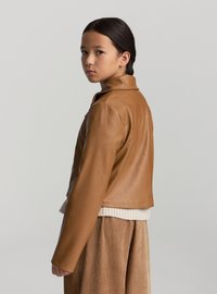 Giacca in pelle marrone con un design leggermente corto, caratterizzata da un colletto a punta e cerniere. Indossata sopra un maglione crema e una gonna beige.