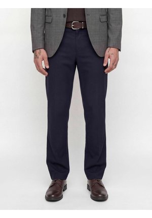Mann trägt marineblaue Hose, braunen Gürtel, grauen karierte Blazer, braune Schuhe, mit tätowierten Händen an den Seiten sichtbar.