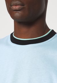 Chemise légère bleu clair à manches courtes avec un col noir côtelé et une touche de vert menthe le long de l'encolure. Texture douce, coupe décontractée.