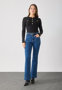 Crni rebrasti longsleeve top s tri zlatna dugmeta; kombiniran s plavim flared trapericama koje imaju stražnje džepove i podvrnute nogavice, nosi se s crnim čizmama.
