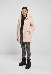 Cappotto con cappuccio in pelliccia sintetica rosa chiaro, con tasche laterali, indossato sopra leggings neri a fantasia e stivali neri caldi. Tessuto morbido, vestibilità rilassata.