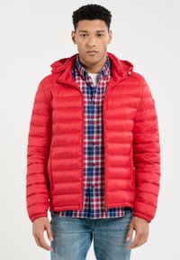 Rød puderjakke med vandret syninger, hætte og lynlåslommer, båret over en blå og rød ternet skjorte og parret med jeans.