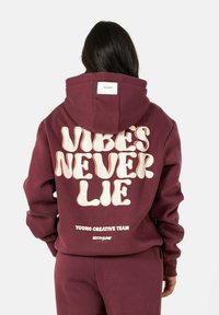 Bordeaux hoodie met grote witte grafische tekst "VIBES NEVER LIE" op de achterkant. Zacht materiaal, kangoeroe-pocket en vaste capuchon.