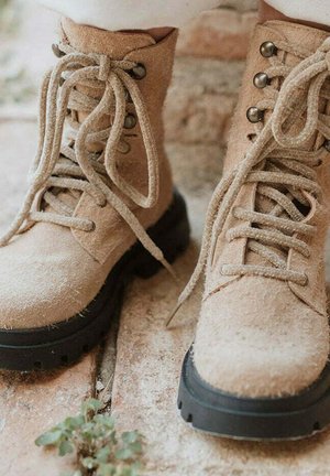 Botas de tobillo de ante color marrón claro con puntas redondas, suelas de goma negra y cordones finos. Cuenta con ojales de metal y una superficie texturizada.