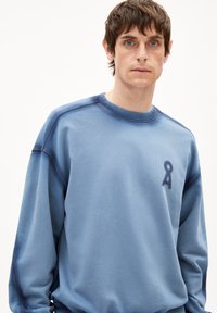 Man met kort bruin haar en blauwe ogen die een losse lichtblauwe sweatshirt draagt met een klein donkerblauw logo op de borst.