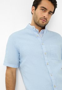 Camicia azzurra a maniche corte realizzata in un tessuto strutturato. Presenta un colletto button-down, una tasca frontale e un rivestimento interno a fantasia floreale.