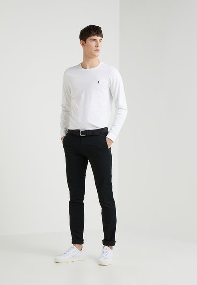 Fehér, hosszú ujjú ing kis logóval, fekete, slim-fit nadrág, fekete öv és fehér sportcipő, egy semleges háttér előtt áll.