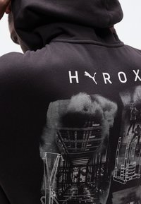 Personne portant un sweat à capuche noir avec le texte "H PUMA ROX" et des graphiques monochromes d'équipement de gym sur le dos.