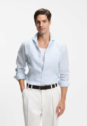 Mężczyzna w jasnoniebieskiej koszuli w paski na białej podkoszulce, białe spodnie z czarnym paskiem, stoi z jedną ręką w kieszeni na białym tle.