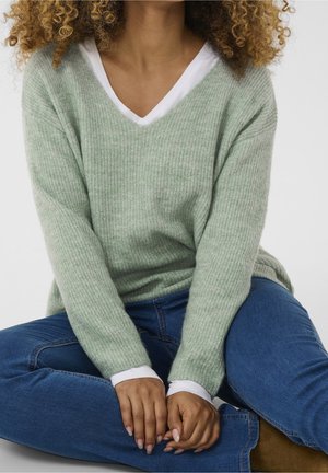 Person med krøllet hår, der sidder med korslagte ben iført en lysegrøn sweater over en hvid skjorte og blå jeans, hænderne hvilende på knæene.