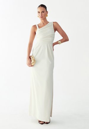 Vrouw in een mouwloze witte jurk tot op de grond, met een gouden clutch, gouden oorbellen en armbanden, poserend tegen een witte achtergrond.