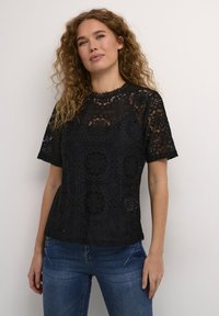 Black lace top featuring a geometric pattern, short sleeves, and a scalloped neckline. Paired with blue jeans, highlighting a relaxed fit.