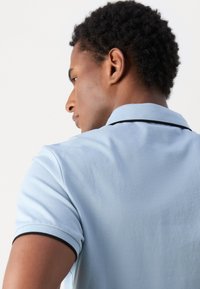 Camisa polo azul claro con acentos de color negro en el collar, de mangas cortas, tejido texturizado y un diseño ajustado. La vista es desde el lado y la parte trasera.