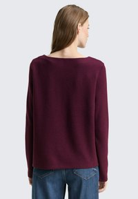 Bordeaux gebreide trui met een brede halslijn en lange mouwen, met een gestructureerde afwerking. De achterkant is eenvoudig en onversierd, gecombineerd met blauwe jeans.