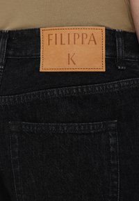Czarne dżinsy z denimu z brązową skórzaną naszywką marki "FILIPPA K." Tkanina ma teksturowaną powierzchnię z widocznymi szwami i tylną kieszenią.