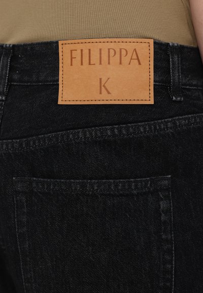 Jeans de mezclilla negra con un parche de cuero marrón claro etiquetado como "FILIPPA K." La tela tiene textura con costuras visibles y un bolsillo trasero.