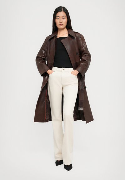 Mujer de pie con las manos en los bolsillos, vistiendo un abrigo de cuero marrón oscuro, una blusa negra, pantalones blancos y zapatos de punta negros, contra un fondo blanco.