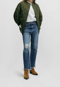 Olivgrön vadderad jacka, vit texturerad blus, slitna blå jeans och bruna klackboots med svart accent vid tå.