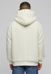 White fleece hoodie with a textured finish, hood, and ribbed cuffs; worn over blue jeans, showing a relaxed fit and soft material.