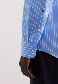 Camicia azzurro chiaro con strisce bianche verticali, polsini con bottoni e un piccolo logo sulla manica. Tessuto liscio con una vestibilità aderente.