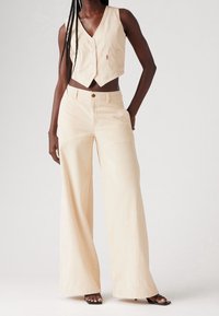 Tummaihottuinen henkilö pukeutunut vaaleanruskeaan kauluspaitatiiviiseen liiviin ja siihen sopiviin leveisiin housuihin, jotka ovat yhdistetty mustiin avonaisiin korkokenkiin.