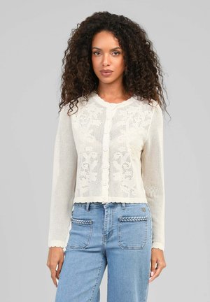 Femme aux cheveux bouclés portant un cardigan en maille crème boutonné avec des motifs floraux et un jean bleu clair avec des détails tressés sur les poches.