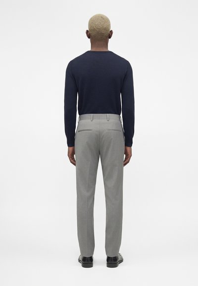 Άνδρας με κοντά ξανθά μαλλιά που φοράει μπλε σκούρο πουκάμισο με μακριά μανίκια, γκρι παντελόνι και μαύρα επίσημα παπούτσια, στέκεται με την πλάτη προς ένα απλό φόντο.