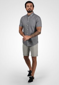 Chemise grise à manches courtes avec un col, dotée d'une poche poitrine, associée à un short gris clair et des baskets noires avec des accents dorés.