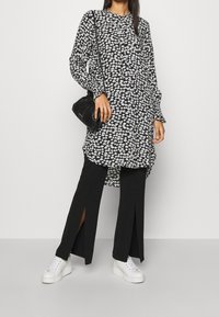Túnica floral en blanco y negro con mangas largas sobre pantalones negros con abertura y zapatillas blancas. Presenta un corte holgado y cuello redondo.