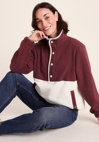 Woman sitting cross-legged wearing a burgundy and white fleece pullover with snap buttons and blue jeans, smiling at the camera.