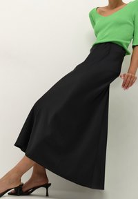 Schwarzer Maxirock aus glattem Stoff mit weitem Schnitt. Kombiniert mit einem eng anliegenden, kurzärmeligen grünen Oberteil. Schlanke, schwarze Peep-Toe-Absätze.