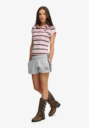 Młoda kobieta ubrana w różową koszulkę polo w paski, szare sportowe szorty z bocznymi paskami oraz brązowe kozaki za kostkę z klamrami, stojąca z rękami w kieszeniach.