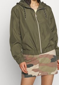 Giacca leggera verde oliva con cappuccio, polsini ed orlo elastici, dotata di zip frontale e due tasche laterali, indossata sopra una gonna a fantasia mimetica.