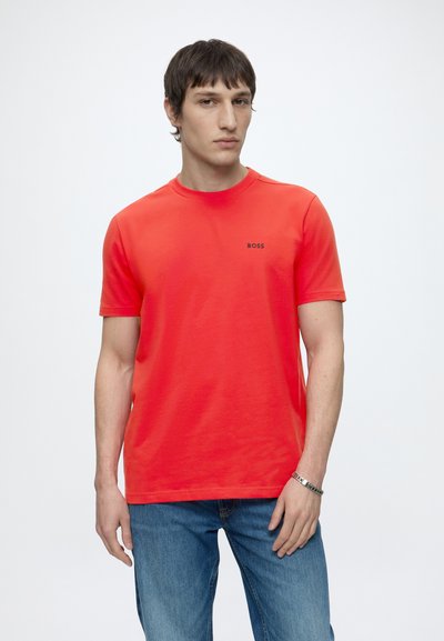 Homme portant un t-shirt BOSS rouge vif et un jean bleu, debout devant un fond blanc uni avec une expression neutre.