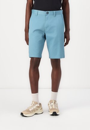 Shorts bleu clair avec un devant plat, fermeture à bouton et poches latérales. Portés avec des chaussures de sport beiges et des chaussettes légères.