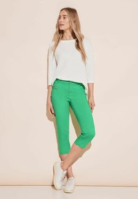 Pantaloni capri verdi realizzati in tessuto morbido, con chiusura a bottone e cerniera, e un accento con tasca laterale. Abbinati a un maglione bianco.