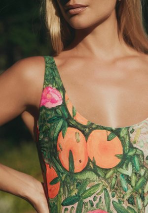 Frau mit blondem Haar, die ein ärmelloses Kleid mit einem grünen und orangefarbenen Blumen- und Fruchtmuster trägt und im Freien im Sonnenlicht steht.