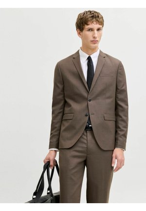 Giovane uomo in abito marrone che tiene una borsa di pelle nera, indossa una camicia bianca e una cravatta nera, in piedi contro uno sfondo chiaro uniforme.