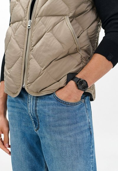 Osoba ubrana w niebieskie dżinsy i beżową pikowaną kamizelkę, z czarną koszulą z długim rękawem i czarnym zegarkiem na nadgarstku, z ręką w kieszeni.