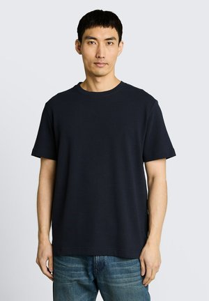 Man draagt een eenvoudig zwart T-shirt met korte mouwen en blauwe spijkerbroek, staat tegen een lichtgrijze achtergrond en kijkt recht vooruit met een neutrale gezichtsuitdrukking.