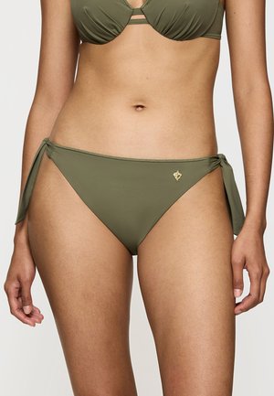 Frau trägt olivgrüne Bikinihose mit seitlichen Bändern und passendem Oberteil, steht vor einem schlichten hellen Hintergrund.