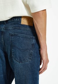 Los vaqueros de mezclilla azul oscuro cuentan con un bolsillo trasero y una etiqueta de cuero marrón con una marca en relieve. La tela tiene una textura desgastada.