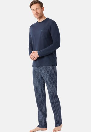 Man standing barefoot wearing a navy long-sleeve shirt with chest pocket and patterned blue pajama pants on a plain light background.