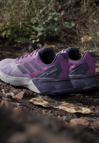 Lilla og grå Adidas Terrex trail running sko på en skovbund med tørre blade og jord.