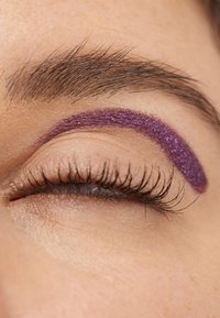Lilla glitter eyeliner er påført over et lukket øye med lange mørke øyevipper, som står i kontrast til hudens lyse tekstur.