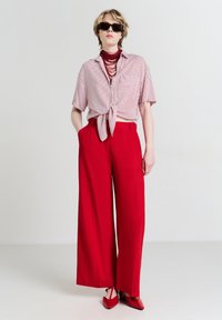 Persona con occhiali da sole neri, collana di perle rosse, camicia a righe con maniche corte annodata in vita, pantaloni larghi rossi e scarpe rosse a punta.