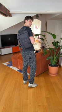 Zwarte rugzak van stof met dubbele gespsluitingen, gedragen door een persoon in een zwart shirt en een broek met een patroon, staand in een kamer met planten.