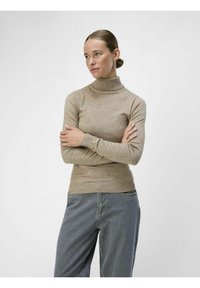 Beżowy sweter w golfa wykonany z dzianiny, z prążkowanymi mankietami i dołem. Połączony z jasnoniebieskimi dżinsami o luźnym fasonie.